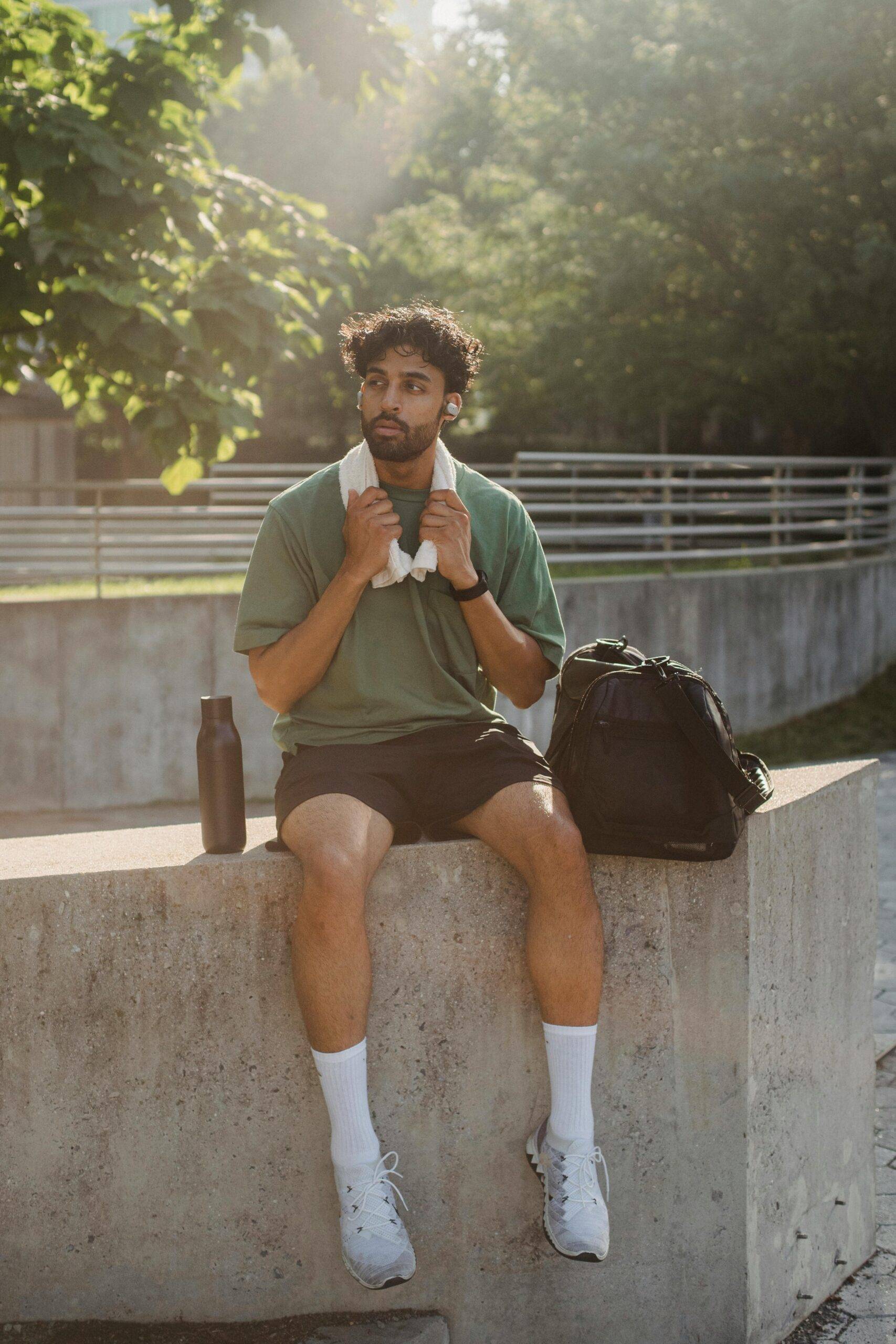 A man in breathable activewear fabric sits on a concrete ledge post-workout, wearing a green performance T-shirt and black athletic shorts. A towel rests around his neck, with a black backpack and reusable water bottle beside him, highlighting the comfort and functionality of UV-protective and antibacterial fabrics in everyday activewear.